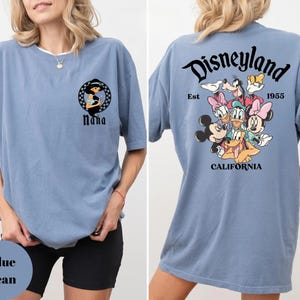 May include: A blue jean colored t-shirt featuring a front graphic of a woman in a black and white circular design with the word "Nana" below. The back of the shirt has a Disneyland graphic with cartoon characters and the text "Disneyland California 1955".