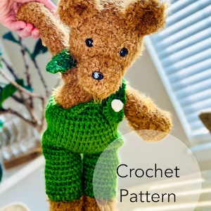 May include: Crocheted brown teddy bear wearing green overalls with a white button. The bear is holding its left arm up. The text "Crochet Pattern" is visible in the image.