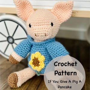 May include: A crocheted pig doll with a light pink body, large ears, and black eyes. It wears a blue sweater with a yellow sunflower applique. The text "Crochet Pattern If You Give A Pig A Pancake" is on the image.