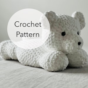 May include: White crocheted stuffed bear toy lying on a white fabric surface. The bear has black eyes and a black nose. The text "Crochet Pattern" is in a white circle on the image.