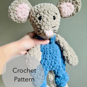 May include: Crochet pattern for a gray and blue plush mouse toy. The mouse has pink ears and a pink nose. The text "Crochet Pattern" is visible in the image.