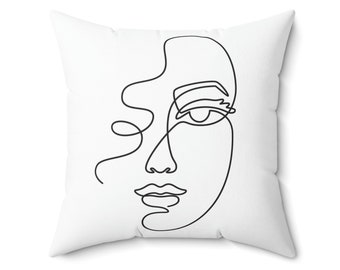 May include: A white throw pillow with a black line drawing of a woman's face. The drawing is in a minimalist style, with only the outline of the face and hair visible.