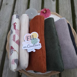 Peut inclure: Plusieurs rouleaux de tissu de couleurs variées, dont blanc, rouille et vert olive, sont disposés dans un panier tressé. Un rouleau présente un motif floral. L'image contient un autocollant avec le texte "FIL en Couleur".