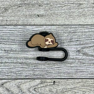 Könnte beinhalten: Ein schwarzer und brauner Faultier-förmiger Haken mit einem schwarzen Metallhaken. Das Faultier schläft mit gekreuzten Armen.