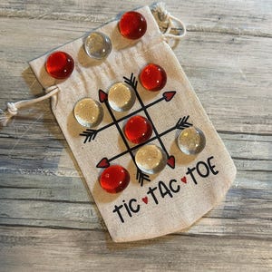 May include: A Tic-Tac-Toe game set with a beige drawstring bag. The bag features a black grid with arrow accents and the words "Tic Tac Toe" in black. The game pieces are red and clear glass gems.