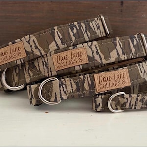 May include: A stack of dog collars with a camouflage pattern in shades of green, brown, and tan. Each collar has a metal D-ring and a leather patch that reads "Dixie Lane Collars".