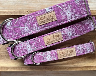Puede incluir: Tres collares para perros con un estampado floral en tonos morados y blancos. Cada collar tiene una hebilla de metal plateado y una pequeña etiqueta rectangular de cuero que dice "Dixie Lane COLLARS". Los collares están colocados sobre una superficie de madera.