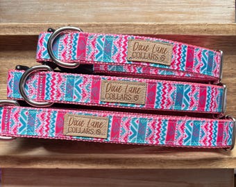 May include: Three dog collars with a pink, teal, and white geometric pattern. Each collar has a silver metal buckle and a brown leather tag that reads "Dixie Lane COLLARS". The collars are stacked on a wooden shelf.