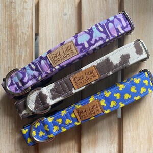 May include: Three dog collars with different patterns. One is purple with mushroom designs, another is brown and white with a cow print, and the last is blue with yellow duck patterns. Each collar has a metal buckle and a leather tag that reads "Dixie Lane Collars."