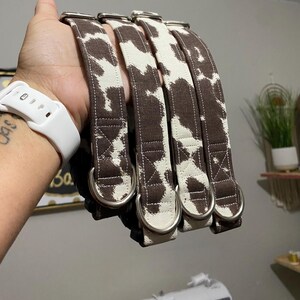 May include: Four dog collars with a brown and white cow print pattern. Each collar has a silver metal D-ring and a matching brown fabric loop. The collars are made of a sturdy fabric and are designed for durability.