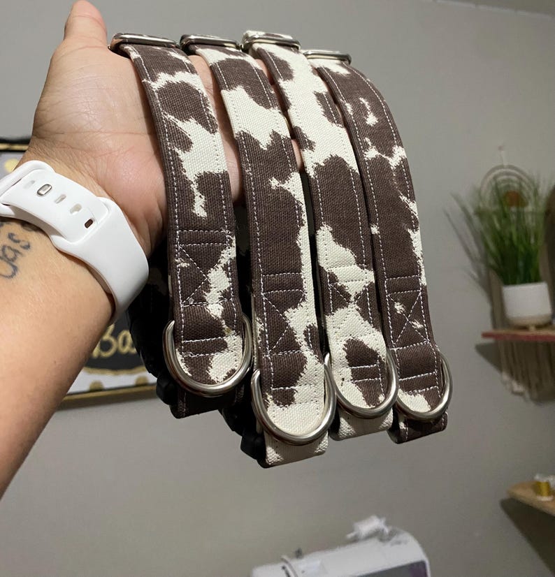 May include: Four dog collars with a brown and white cow print pattern. Each collar has a silver metal D-ring and a matching brown fabric loop. The collars are made of a sturdy fabric and are designed for durability.