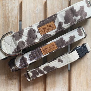May include: Three dog collars with a brown and white cow print pattern. The collars have metal hardware and a leather tag that reads "Dixie Lane Collars". The collars are of different sizes.