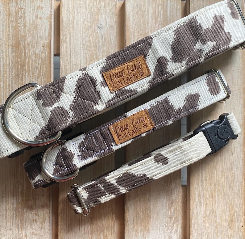 May include: Three dog collars with a brown and white cow print pattern. The collars have metal hardware and a leather tag that reads "Dixie Lane Collars". The collars are of different sizes.
