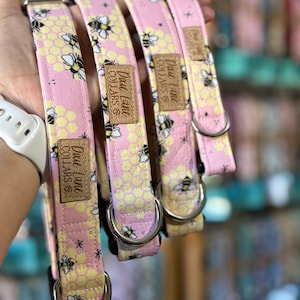 May include: Pink dog collars featuring a bee and honeycomb design. Each collar has a silver metal D-ring and a brown leather tag with "Dune Lane Collars". The collars are made of fabric and have a black and yellow bee design.