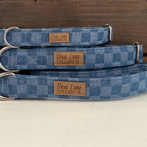 May include: Three blue denim dog collars with a checkered pattern are stacked on top of each other. Each collar has a silver metal D-ring and a brown leather tag that reads "Dixie Lane Collars". The collars are displayed on a white surface.