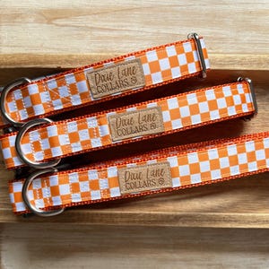 May include: Three orange and white checkered dog collars with silver hardware are displayed on a wooden surface. Each collar has a brown patch with the text "Dixie Lane COLLARS". The collars are arranged in a row, showcasing their design and features.