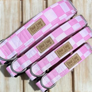 Może przedstawiać: Cztery różowo-białe obroże dla psów w kratę o różnych rozmiarach. Każda obroża ma srebrny D-ring i brązową skórzaną naszywkę z napisem "Dixie Lane Collars". Obroże są ułożone po przekątnej.