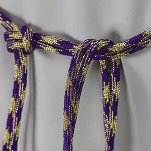 Op de afbeelding: Close-up van een decoratief paars en goudkleurig koord, in het midden geknoopt. Het koord heeft een geweven ontwerp met een herhalend patroon van gouden accenten tegen een paarse achtergrond. Het koord hangt tegen een lichtgrijze achtergrond.