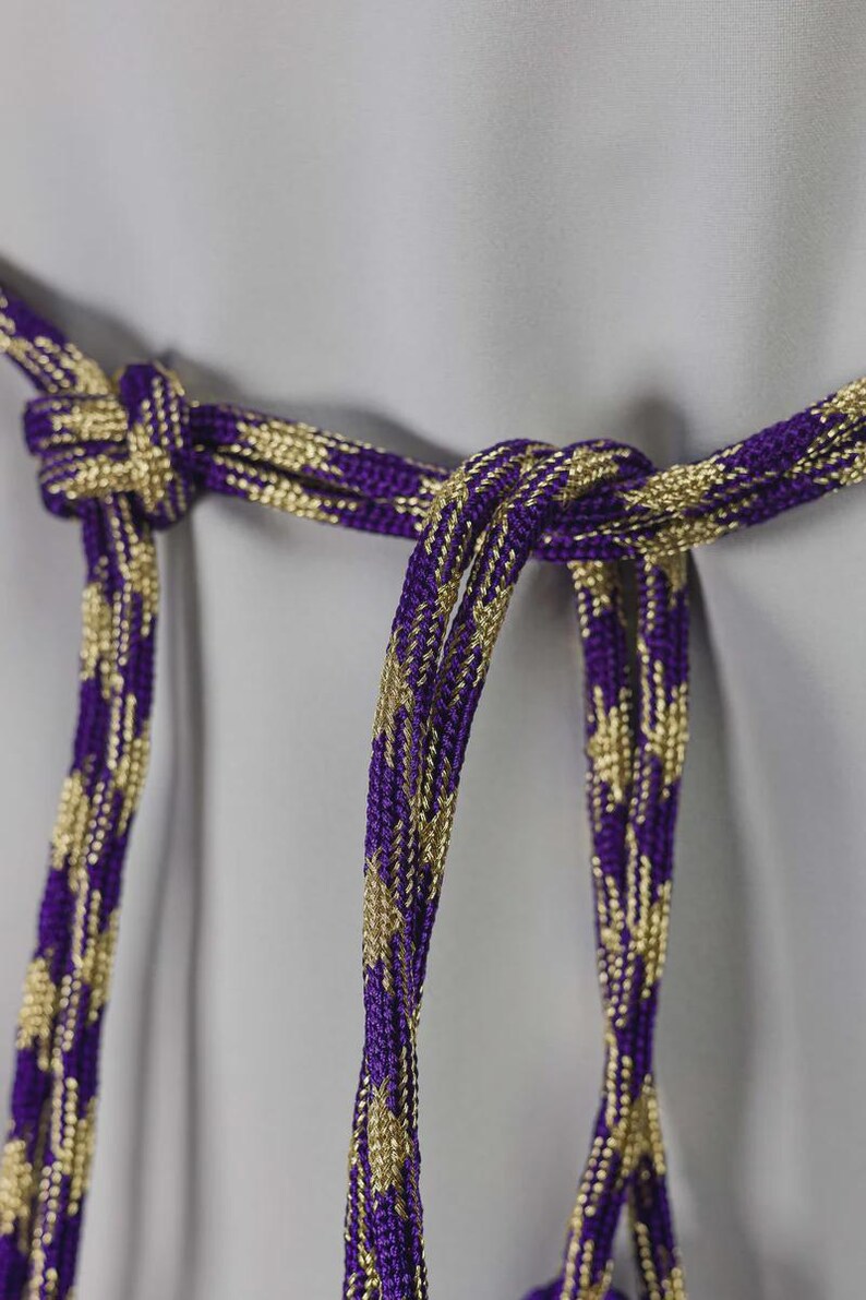 Op de afbeelding: Close-up van een decoratief paars en goudkleurig koord, in het midden geknoopt. Het koord heeft een geweven ontwerp met een herhalend patroon van gouden accenten tegen een paarse achtergrond. Het koord hangt tegen een lichtgrijze achtergrond.