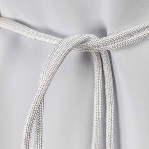 Op de afbeelding: Close-up van een zilverwit touw dat in een knoop is gelegd. Het touw is dik en getextureerd, met een geweven uiterlijk. De achtergrond is effen lichtgrijs en biedt een neutrale achtergrond voor het touw.