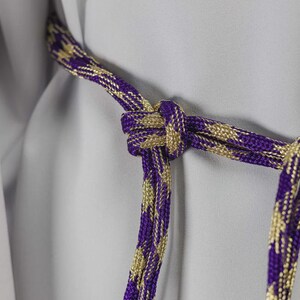 Op de afbeelding: Close-up van een decoratief koord met een knoop. Het koord is een combinatie van paars en goud, met een geweven patroon. De achtergrond is een lichtgrijze stof. Het koord wordt waarschijnlijk gebruikt voor decoratieve doeleinden.