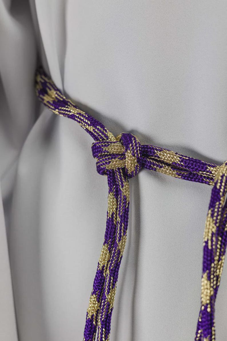 Op de afbeelding: Close-up van een decoratief koord met een knoop. Het koord is een combinatie van paars en goud, met een geweven patroon. De achtergrond is een lichtgrijze stof. Het koord wordt waarschijnlijk gebruikt voor decoratieve doeleinden.