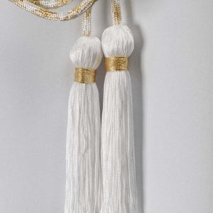 Op de afbeelding: Twee witte kwasten met gouden accenten hangen tegen een witte achtergrond. De kwasten zijn gemaakt van een zijdeachtig, getextureerd materiaal en zijn versierd met gouden banden. Het koord boven de kwasten is wit en goud.