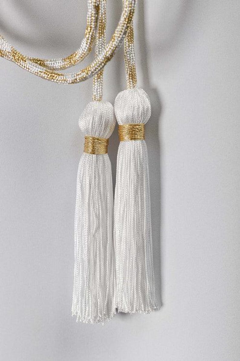 Op de afbeelding: Twee witte kwasten met gouden accenten hangen tegen een witte achtergrond. De kwasten zijn gemaakt van een zijdeachtig, getextureerd materiaal en zijn versierd met gouden banden. Het koord boven de kwasten is wit en goud.