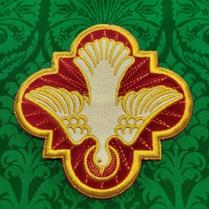 以下が含まれることがあります： 中央に白い鳩が描かれた、金と赤の刺繍パッチ。鳩は羽を広げて前を向いています。パッチはハート型です。