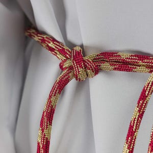 Op de afbeelding: Close-up van een decoratief rood en goudkleurig touw dat in een knoop is gelegd. Het touw heeft een herhalend goudkleurig patroon tegen een rode achtergrond. De achtergrond is een lichtgrijze stof.