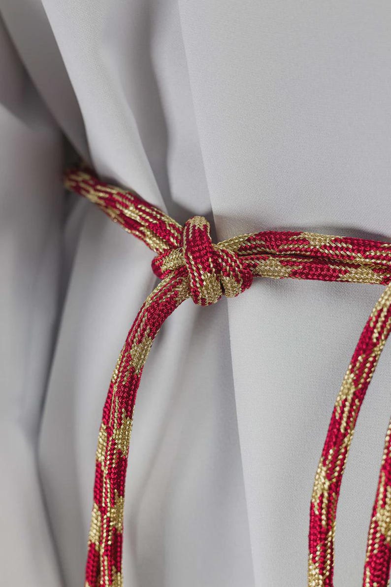 Op de afbeelding: Close-up van een decoratief rood en goudkleurig touw dat in een knoop is gelegd. Het touw heeft een herhalend goudkleurig patroon tegen een rode achtergrond. De achtergrond is een lichtgrijze stof.