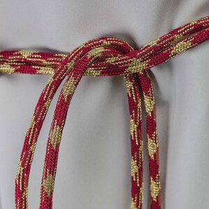 Op de afbeelding: Close-up van een rood en goudkleurig decoratief koord, in het midden geknoopt. Het koord heeft een herhalend patroon van gouden ruiten op een rode achtergrond. Het koord wordt waarschijnlijk gebruikt voor decoratieve doeleinden.