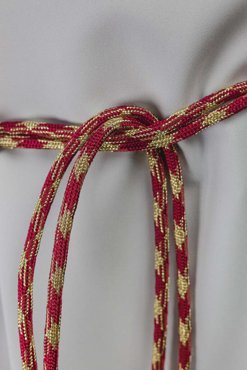 Op de afbeelding: Close-up van een rood en goudkleurig decoratief koord, in het midden geknoopt. Het koord heeft een herhalend patroon van gouden ruiten op een rode achtergrond. Het koord wordt waarschijnlijk gebruikt voor decoratieve doeleinden.