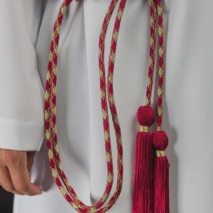 Op de afbeelding: Een rode en gouden touwriem met kwastjes. De riem is gemaakt van een gedraaide rood-gouden koord, geknoopt in de taille. De kwastjes zijn dieprood van kleur en zijn bevestigd aan de uiteinden van de riem.