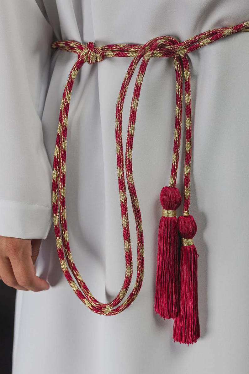 Op de afbeelding: Een rode en gouden touwriem met kwastjes. De riem is gemaakt van een gedraaide rood-gouden koord, geknoopt in de taille. De kwastjes zijn dieprood van kleur en zijn bevestigd aan de uiteinden van de riem.
