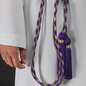 Op de afbeelding: Een paars en goud geweven koord met kwastjes. Het koord is geknoopt en hangt naar beneden, met twee paarse kwastjes aan het uiteinde. Het koord wordt over een wit kledingstuk gedragen.