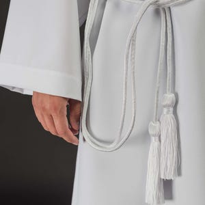 Puede incluir: Túnica litúrgica blanca con cordón y borlas plateadas. La túnica tiene mangas largas y un diseño sencillo. El cordón plateado está atado a la cintura y cuelga con dos borlas. La tela parece ser un material liso y opaco.