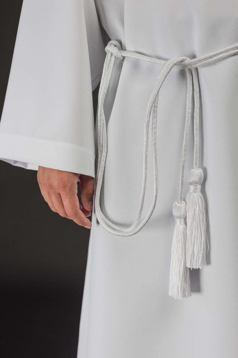Op de afbeelding: Witte liturgische toga met een zilveren koord en kwasten. De toga heeft lange mouwen en een eenvoudig ontwerp. Het zilveren koord is om de taille geknoopt en hangt naar beneden met twee kwasten. De stof lijkt een glad, ondoorzichtig materiaal te zijn.