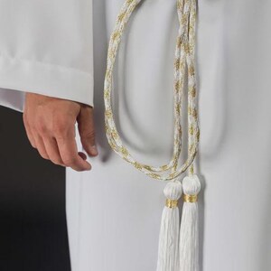 Op de afbeelding: Een wit en goud gevlochten koordriem met kwastjes. De riem is geknoopt in de taille en heeft een goud en wit gevlochten ontwerp. Twee witte kwastjes met gouden accenten hangen aan de riem.