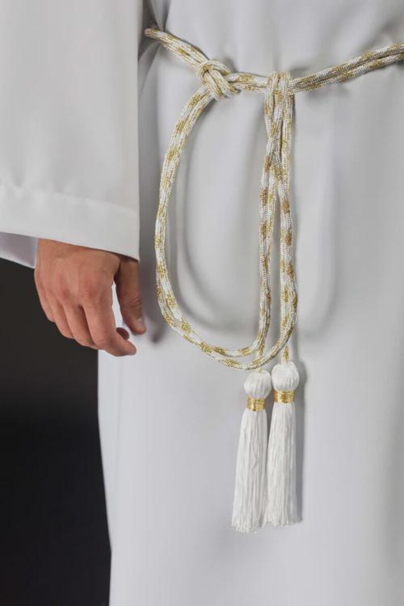 Op de afbeelding: Een wit en goud gevlochten koordriem met kwastjes. De riem is geknoopt in de taille en heeft een goud en wit gevlochten ontwerp. Twee witte kwastjes met gouden accenten hangen aan de riem.