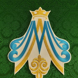 May include: A white embroidered patch with a gold crown and the letter "M" in blue and gold. The patch is shaped like a stylized flame with a gold and blue swirl at the bottom.
