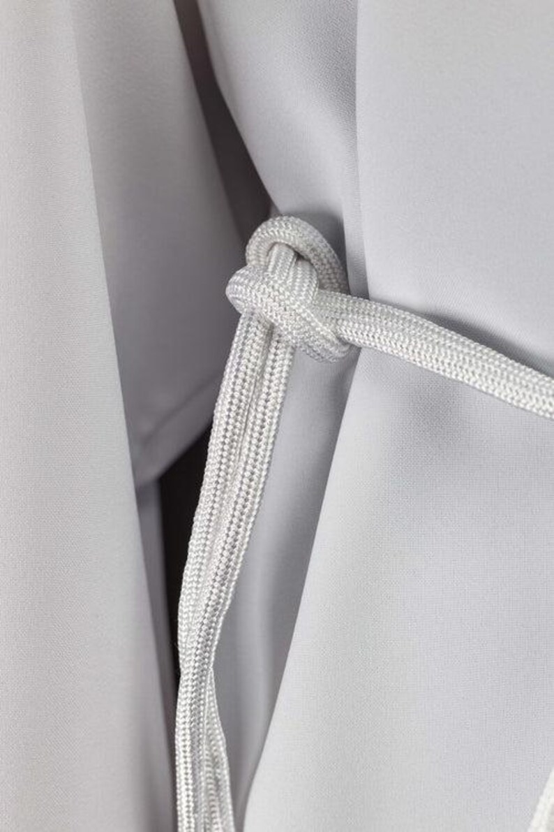 Op de afbeelding: Close-up van een wit touw dat in een knoop is gelegd, met een getextureerd uiterlijk. Het touw is geplaatst tegen een achtergrond van lichtgrijze stof, waardoor een contrast in texturen en tinten ontstaat. De afbeelding benadrukt de ingewikkelde knoop van het touw.