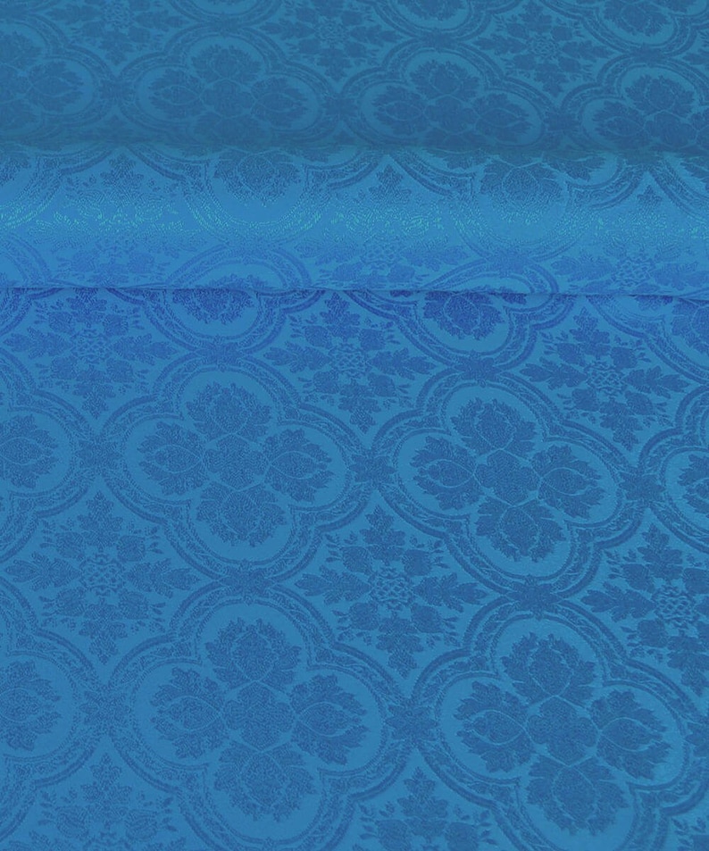 Op de afbeelding: Een close-up van een blauwe stof met een herhalend patroon van gestileerde bloemen en bladeren. De stof heeft een subtiele glans en een textuur.