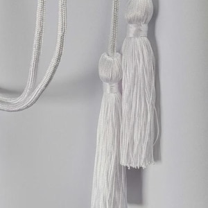 Op de afbeelding: Close-up van witte kwasten en koorden tegen een witte achtergrond. De kwasten zijn gemaakt van vele strengen witte draad en de koorden zijn gevlochten. De kwasten zijn met een decoratieve knoop aan de koorden bevestigd.