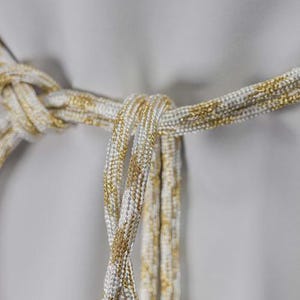 Op de afbeelding: Close-up van een decoratieve riem met een geknoopt ontwerp. De riem is gemaakt van een wit en goudkleurig metallic touwachtig materiaal, met de gouden draden door het wit verweven. De achtergrond is effen grijs.