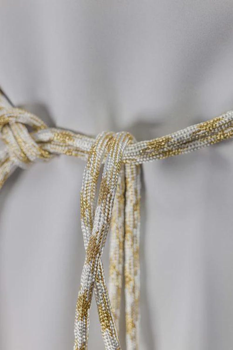 Op de afbeelding: Close-up van een decoratieve riem met een geknoopt ontwerp. De riem is gemaakt van een wit en goudkleurig metallic touwachtig materiaal, met de gouden draden door het wit verweven. De achtergrond is effen grijs.