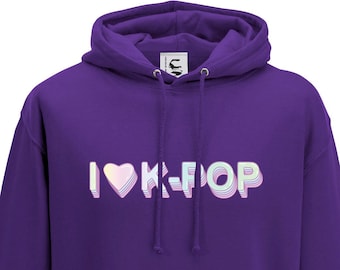 Può includere: Una felpa con cappuccio viola con la scritta "I ♡ K-POP" in un design a strati pastello. La felpa ha una tasca frontale e un cappuccio con coulisse. Il capo è realizzato in un materiale morbido.