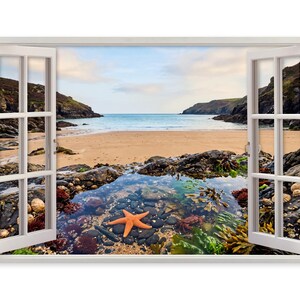 Puede incluir: Un cuadro de pared que muestra un marco de ventana blanco abierto con vistas a una escena costera. La imagen presenta una playa de arena, acantilados rocosos y una poza de marea en primer plano con una estrella de mar naranja, piedras oscuras y algas.