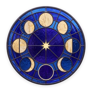 May include: A circular stained glass panel featuring a central eight-pointed star surrounded by a series of moon phases. The design uses textured deep blue glass for the background and lighter cream-coloured glass for the moon shapes, all held together by dark metal lead lines.