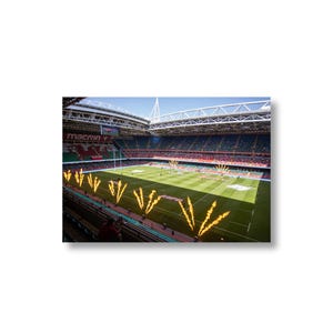 Könnte beinhalten: Ein großes Stadion mit einem grünen Rasen und einem rot-weißen Banner mit der Aufschrift "macron". Es befinden sich viele Menschen auf den Tribünen und einige auf dem Spielfeld. Außerdem schießen mehrere gelbe Flammen aus dem Boden.